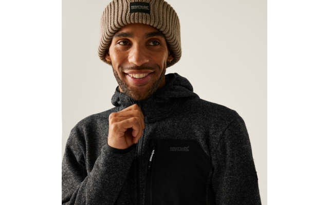Ein Mann mit brauner Beanie-Mütze und schwarzem Pullover lächelt und hält den Kragen seines Pullovers