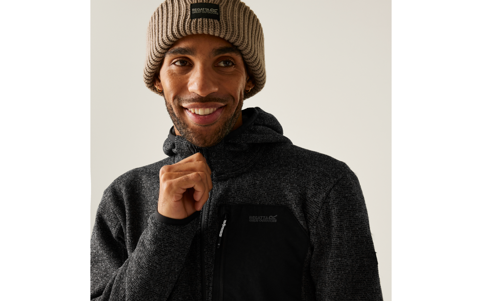 Ein Mann mit brauner Beanie-Mütze und schwarzem Pullover lächelt und hält den Kragen seines Pullovers
