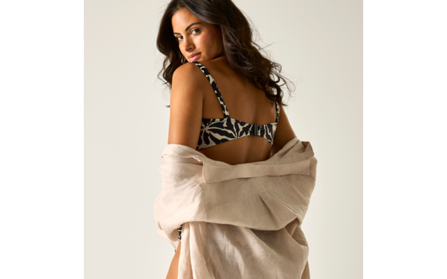 Frau leicht von hinten gesehen, trägt gemusterten BH und offenes beige Leinenhemd über den Schultern, schaut über die Schulter, dunkle lange Haare, heller Hintergrund