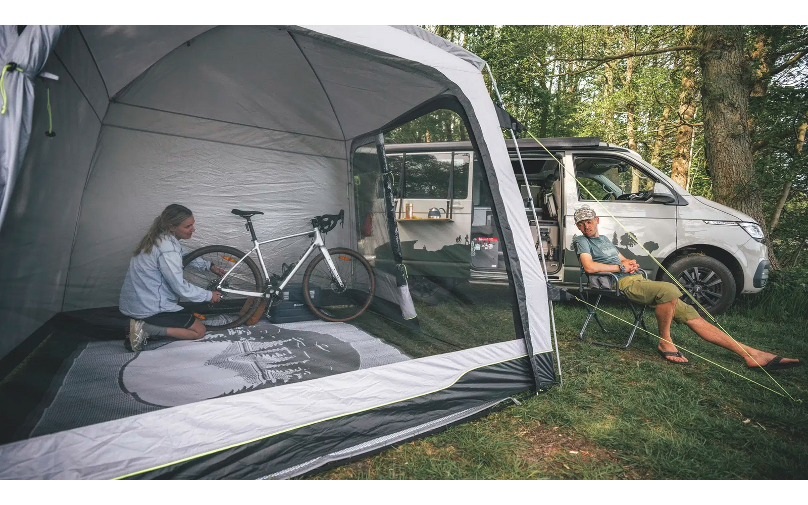 Une personne répare un vélo à l'intérieur d'une tente, une autre personne est assise sur une chaise pliante devant un camping-car