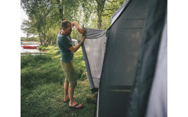 Homme monte une tente grise en plein air, entouré d'arbres et d'herbe