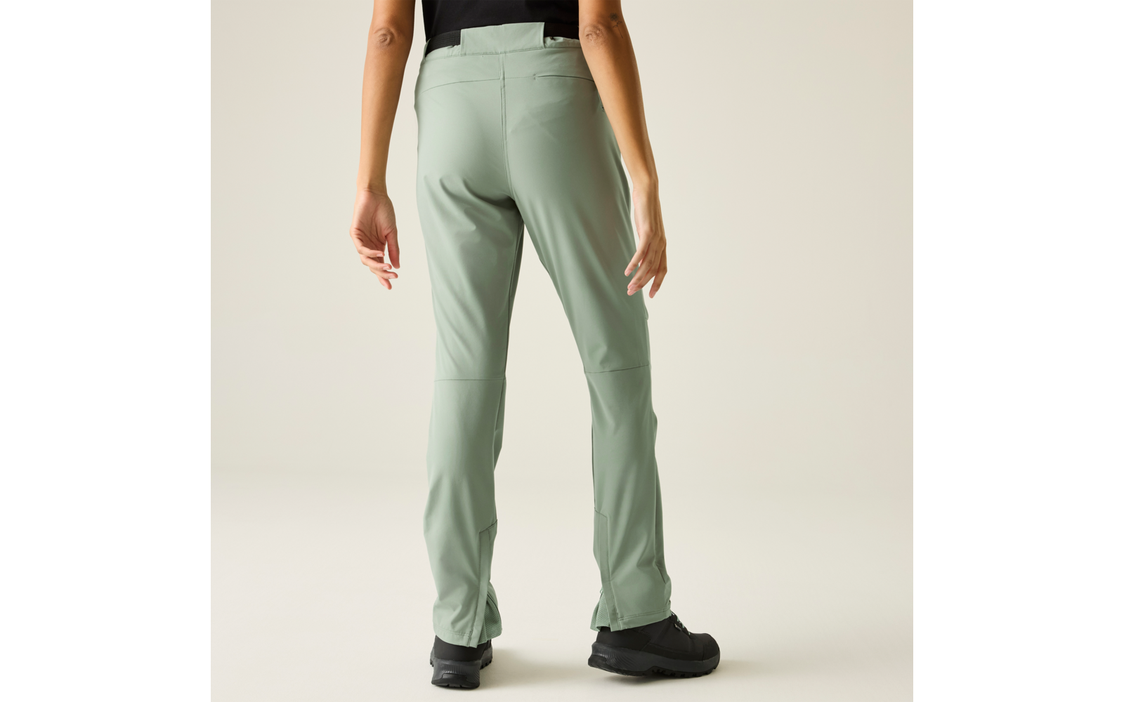 Achteraanzicht van een persoon met lichtgroene broek, zwart bovenstuk en zwarte schoenen, voor een lichte neutrale achtergrond