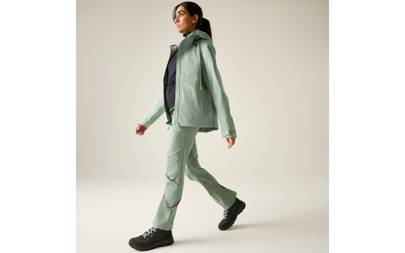 Persoon loopt naar rechts, draagt mintgroene jas en broek, zwart bovenstuk en zwarte schoenen, haar in een paardenstaart, lichte effen achtergrond