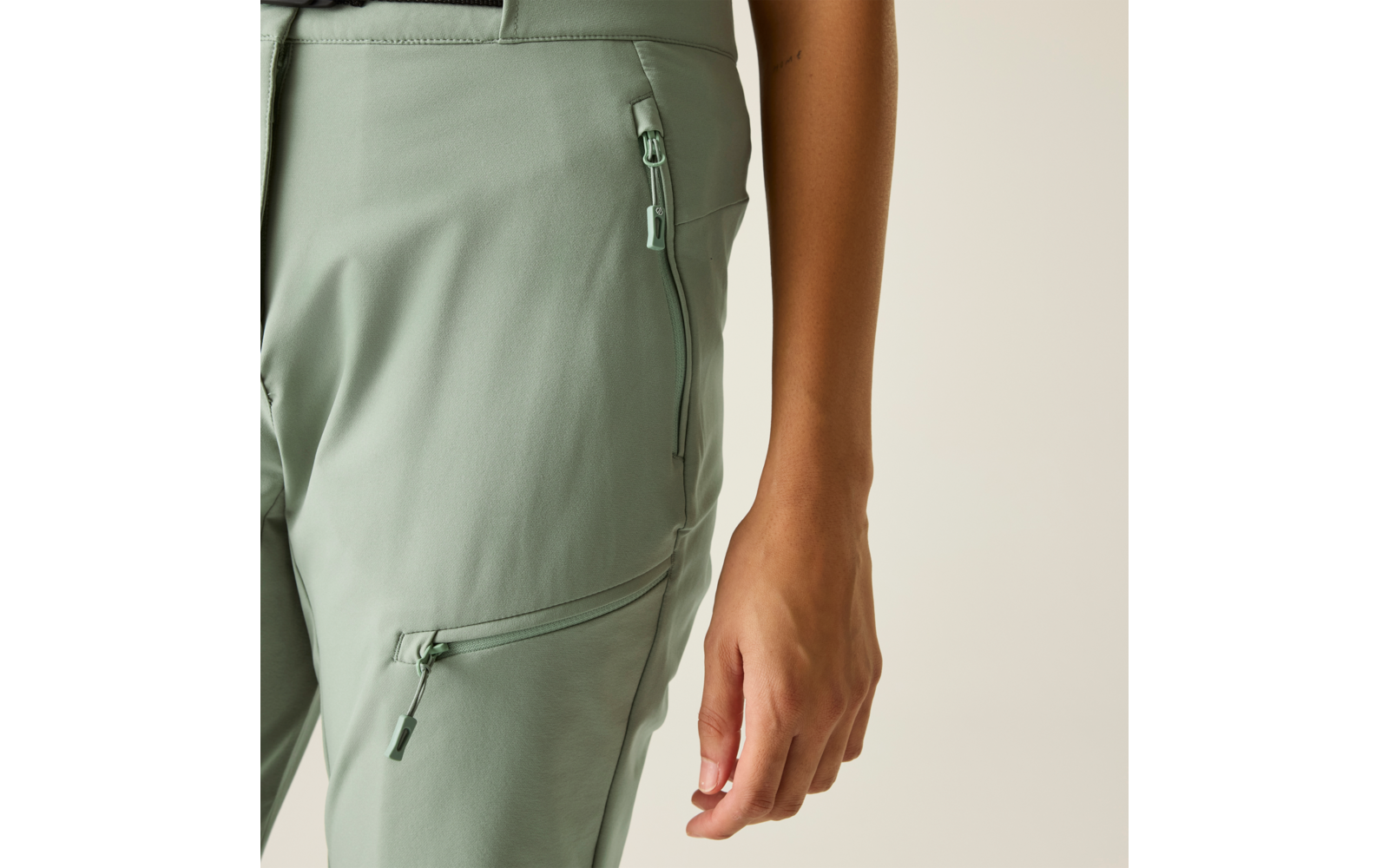 Close-up van heup en dij van een persoon in lichtgroene broek met twee ritszakken, rechter onderarm en hand zichtbaar, beige achtergrond