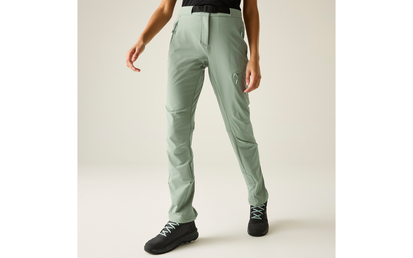 Onderlichaam van een persoon met lichtgroene broek en zwarte schoenen met lichtgroene veters, handen langs het lichaam, neutrale achtergrond