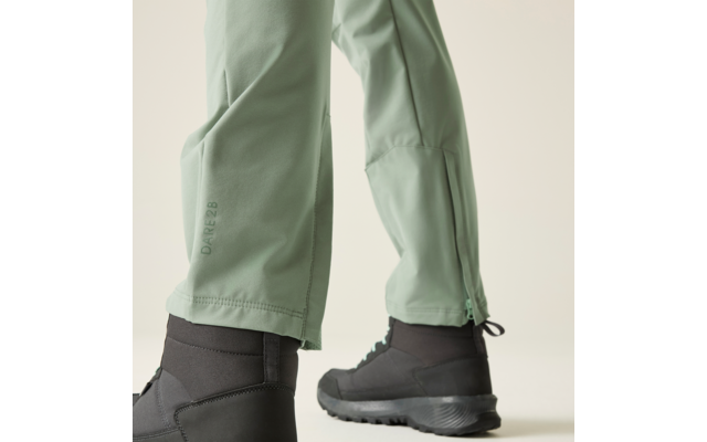 Onderste deel van twee benen in lichtgroene broek met ritssluiting aan de zoom, zichtbare tekst DARE2B en zwarte laarzen op een lichte achtergrond