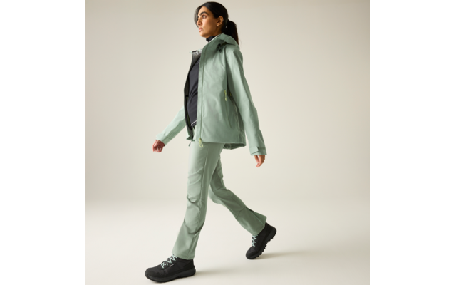 Persoon in lichtgroene jas en bijpassende broek met zwarte wandelschoenen, lopend voor een effen lichte achtergrond