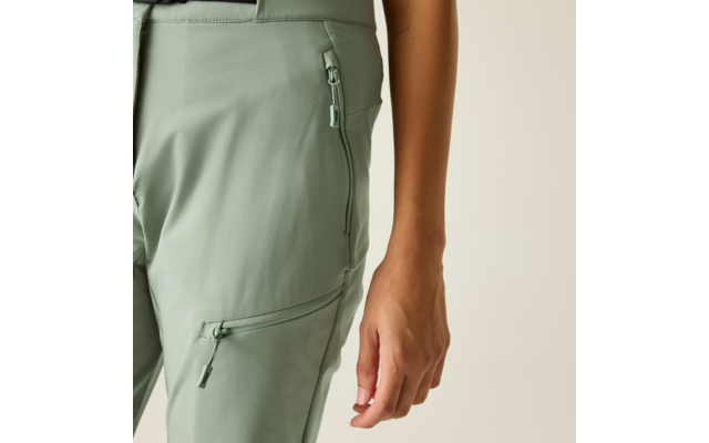 Close-up van heup en dij van een persoon in lichtgroene broek met twee ritszakken, rechter onderarm en hand zichtbaar, beige achtergrond