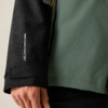 Nahaufnahme einer Hand an einer Jacke: schwarzer Ärmel mit vertikaler weißer Schrift und kleinem weißem Logo, grüner Jackenkörper, sichtbare Reißverschluss-Ziehschlaufe in Gelb