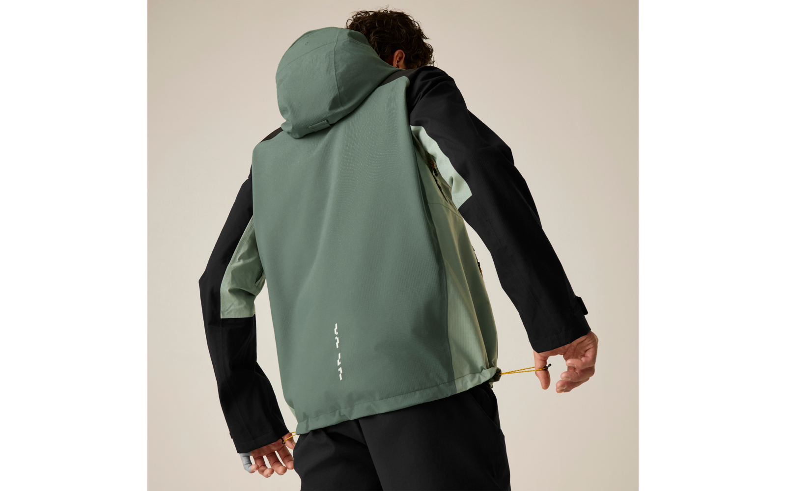 Rückenansicht einer Person mit grüner Kapuzenjacke, schwarzen Ärmeln und hellgrünen Seitenteilen, die am Saum einen gelben Kordelzug hält; schwarze Hose; neutraler Hintergrund