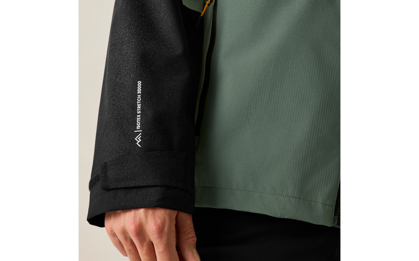 Nahaufnahme einer Hand an einer Jacke: schwarzer Ärmel mit vertikaler weißer Schrift und kleinem weißem Logo, grüner Jackenkörper, sichtbare Reißverschluss-Ziehschlaufe in Gelb