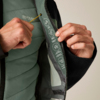 Zwei Hände halten eine schwarze Jacke offen und zeigen das grüne, gesteppte Innenfutter mit weißen aufgedruckten Symbolen, gelbem Kordelzug und Reißverschluss
