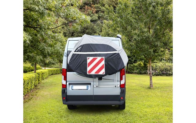Vista posteriore di un camper argento su prato, con una copertura grigio-nera e un pannello rettangolare a strisce diagonali rosse e bianche fissato sul retro, alberi e prato sullo sfondo