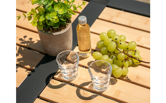 Potplant, klein flesje met gele vloeistof, een tros groene druiven en twee doorzichtige glazen op een tafel van houten latten