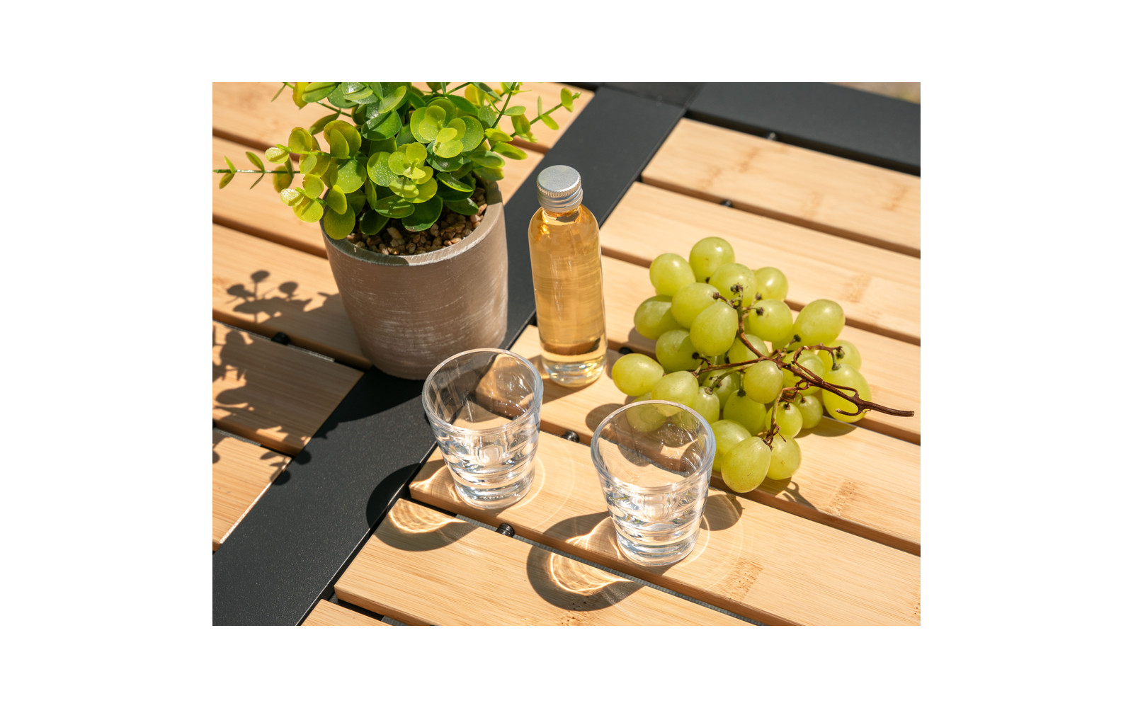 Topfpflanze, kleine Flasche mit gelber Flüssigkeit, eine Traubenrispe und zwei durchsichtige Gläser auf einem Tisch aus Holzlatten