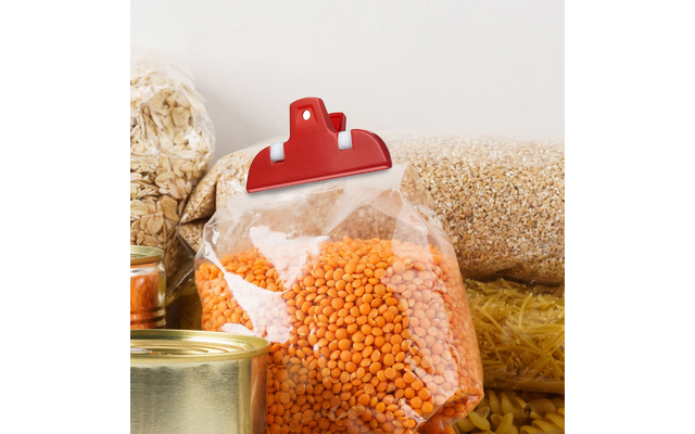 Doorzichtige plastic zak met oranje linzen, bovenop dichtgeklemd met een rode clip; op de achtergrond een blik, pot havermout en graan- en pastaverpakkingen