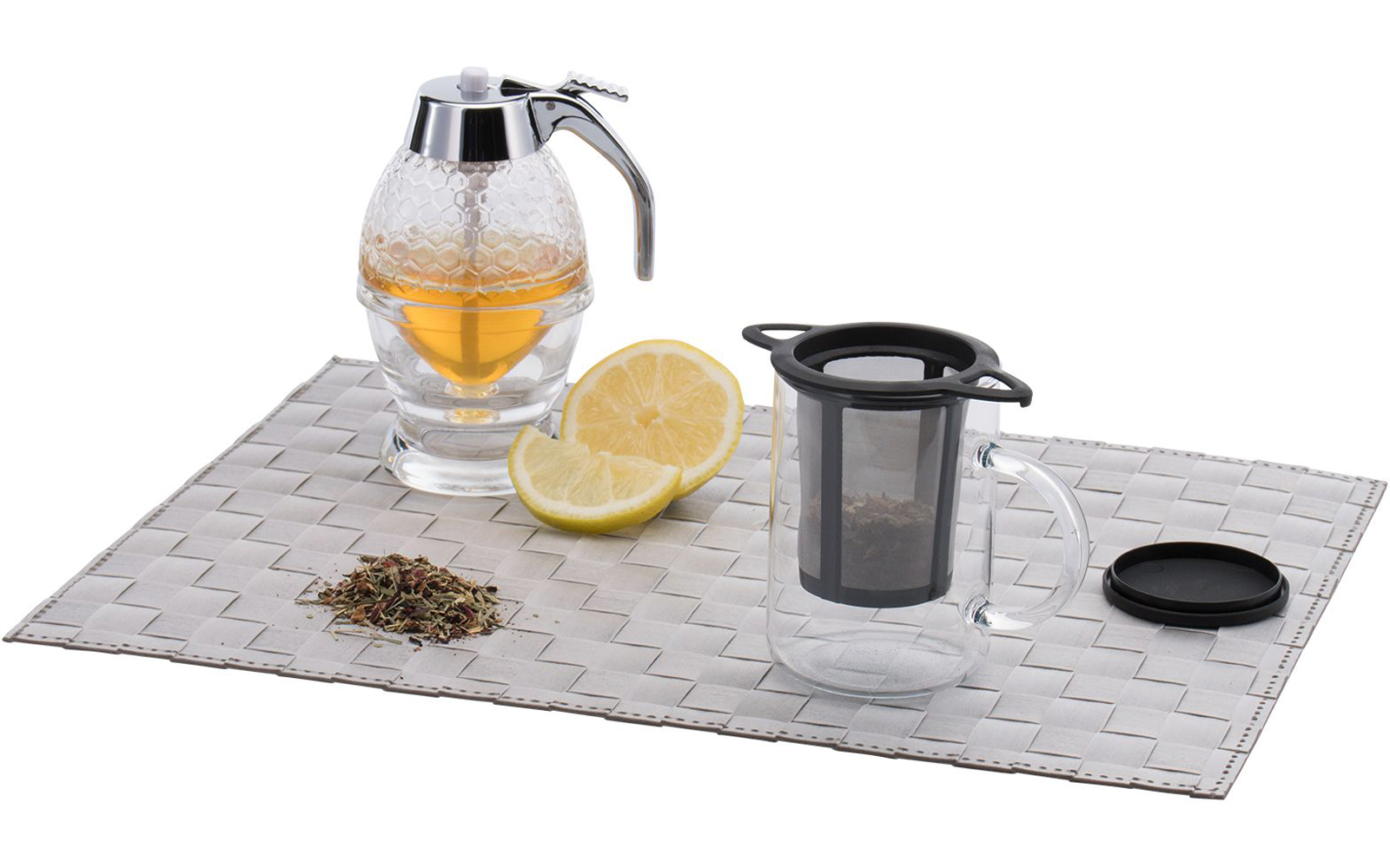 Récipient en verre avec couvercle chromé et liquide jaune, citron coupé en deux, petit tas de feuilles sèches et tasse en verre transparente avec tamis noir sur napperon tressé