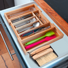 Open kitchen drawer with bamboo organizer holding cutlery, tongs, measuring spoons, two silicone spatulas (green, pink), and small brush