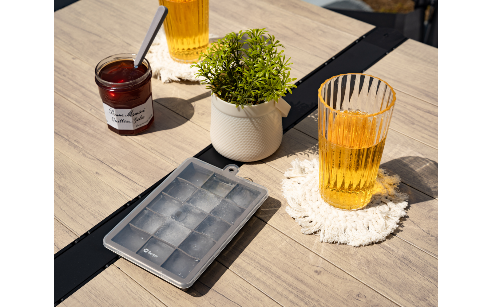 Holztisch mit Eiswürfelschale, zwei gefüllten Gläsern auf gehäkelten Untersetzern, weißem Pflanzenübertopf mit grüner Pflanze und offenem Marmeladenglas