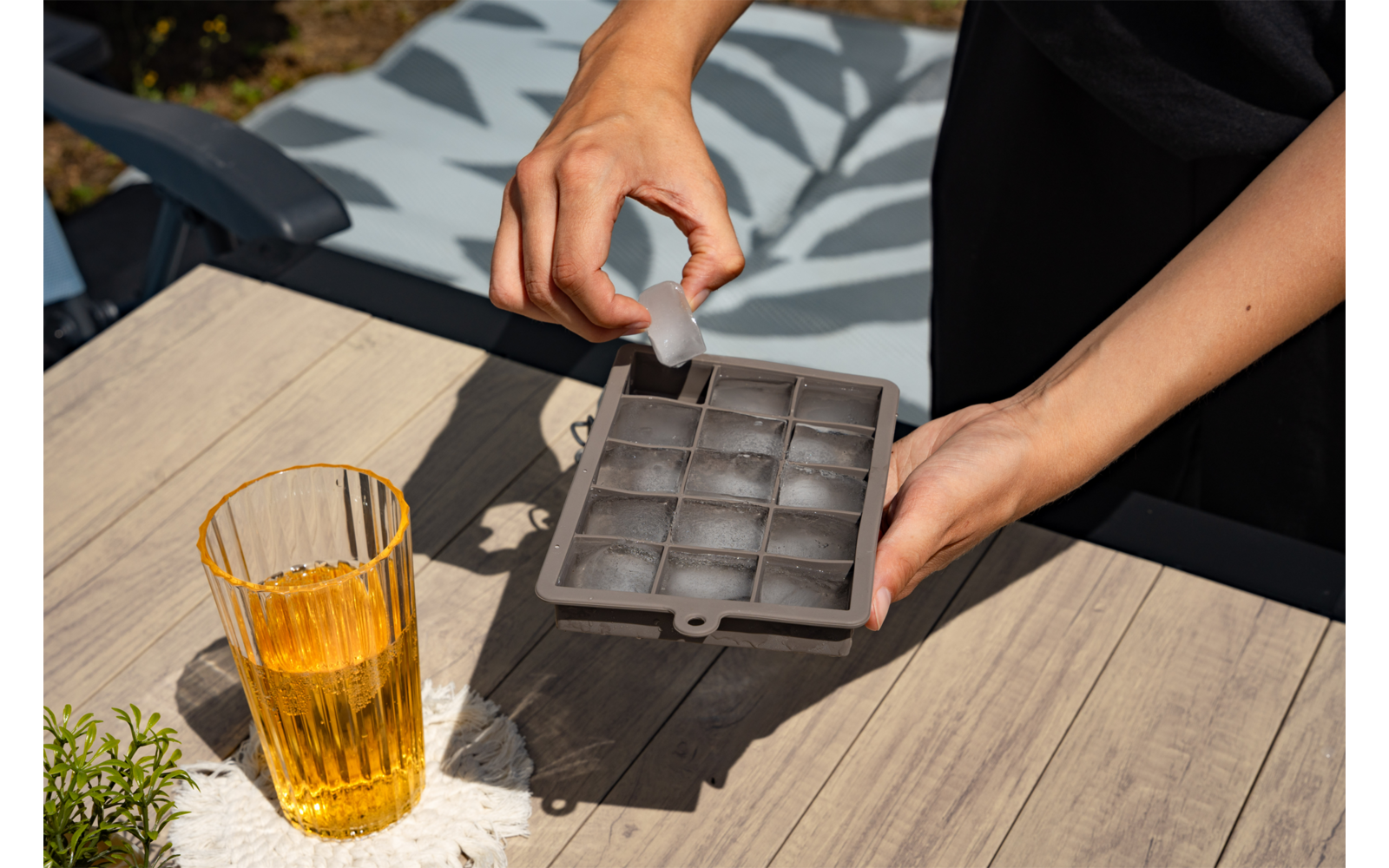 Hände halten eine Eiswürfelform, eine Hand setzt einen Eiswürfel ein; daneben Glas mit brauner Flüssigkeit auf Holztisch, gemustertes Kissen im Hintergrund