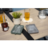 Holztisch mit zwei hohen Gläsern mit bernsteinfarbener Flüssigkeit auf weißen Untersetzern, zwei Eiswürfelformen mit Eis, kleinweißer Blumentopf mit grüner Pflanze, Glasgefäß mit weißen Zuckerwürfeln und Glas mit roter Konfitüre und Löffel, unscharfe