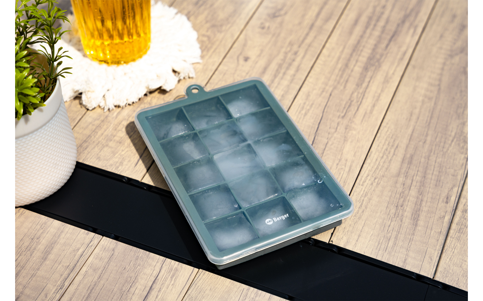 Türkise Eiswürfelform mit gefüllten Fächern auf Holzterrasse, daneben weißer Blumentopf und gelbes Glas auf weißem Untersetzer