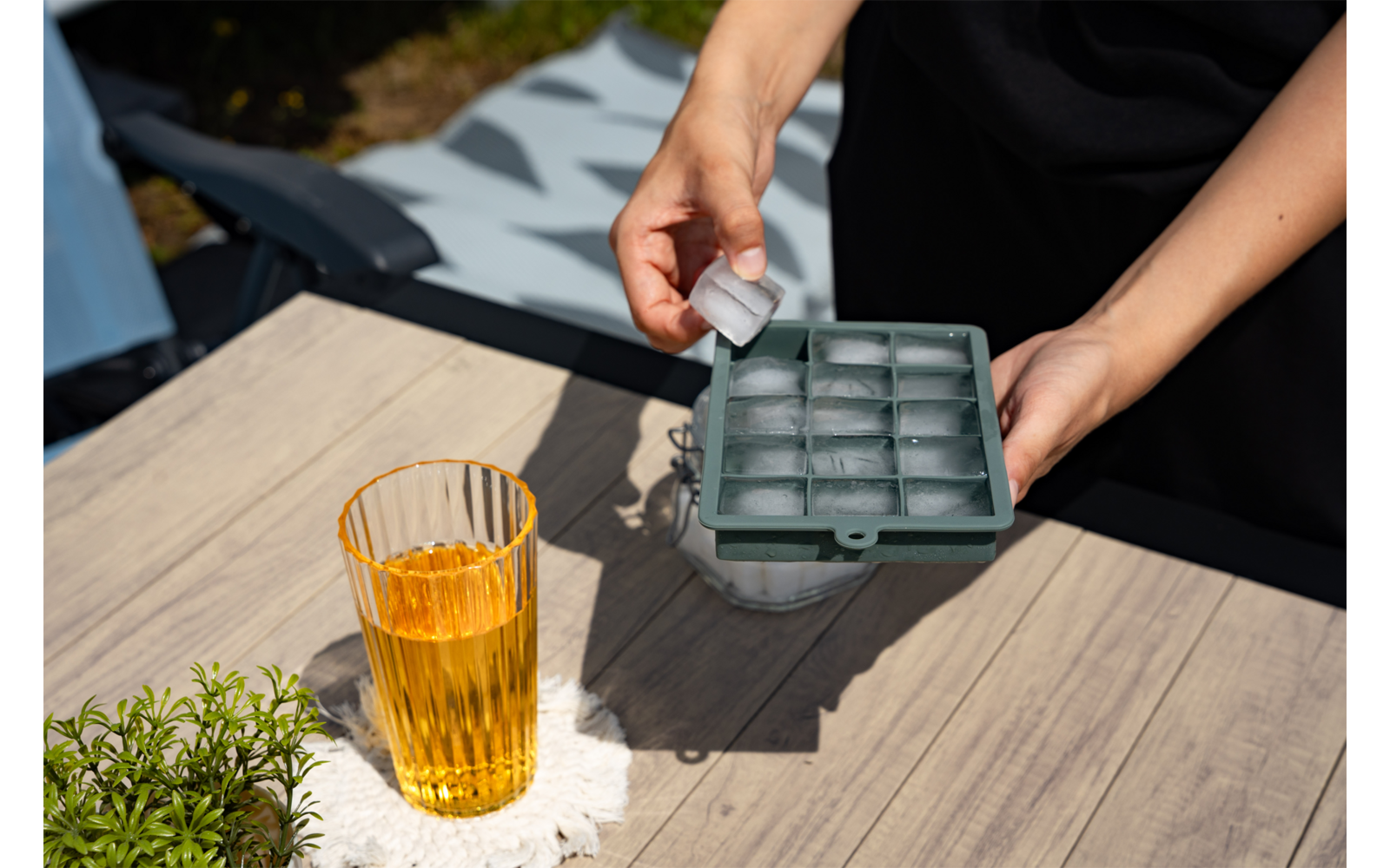 Zwei Hände halten einen Eiswürfel und ein grünes Silikon-Eiswürfelfach über einem Glas mit bernsteinfarbener Flüssigkeit auf einem Holztisch; kleine Topfpflanze links; weißes Tuch unter dem Glas