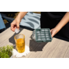Hände halten eine Eiswürfelform und setzen einen Eiswürfel in ein hohes Glas mit bernsteinfarbener Flüssigkeit auf einem Holztisch; Untersetzer und kleiner Topf mit Pflanze daneben