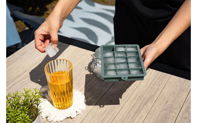 Hände halten eine Eiswürfelform und setzen einen Eiswürfel in ein hohes Glas mit bernsteinfarbener Flüssigkeit auf einem Holztisch; Untersetzer und kleiner Topf mit Pflanze daneben