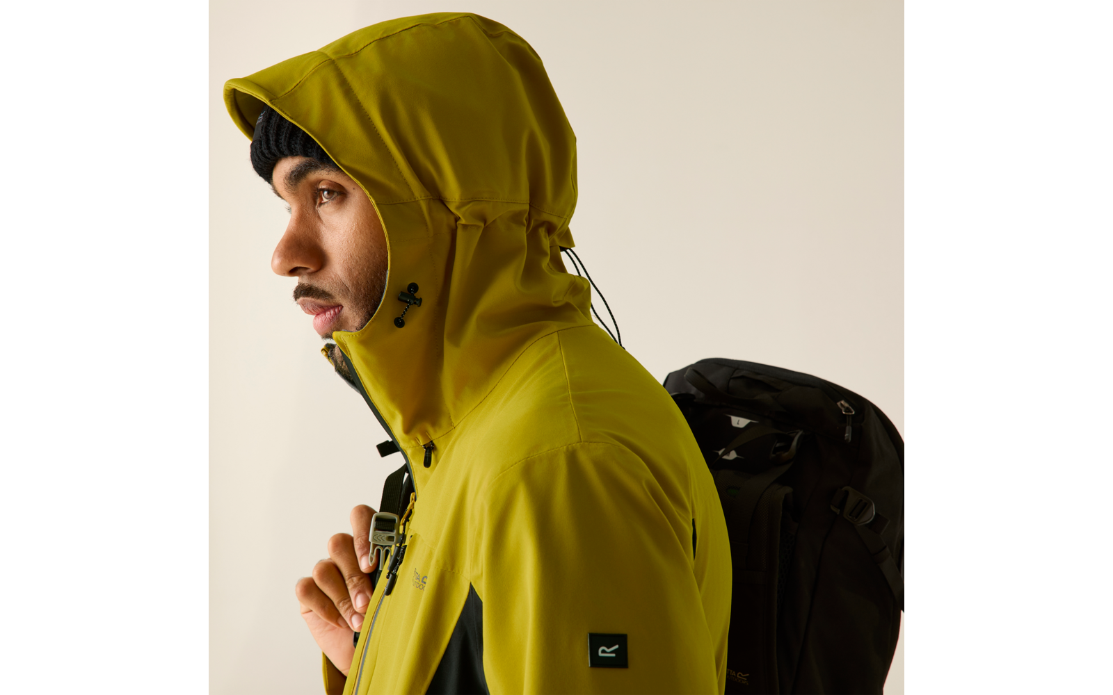 Person im Seitenprofil mit aufgezogener gelbgrüner Kapuzenjacke, schwarzer Mütze und schwarzem Rucksack