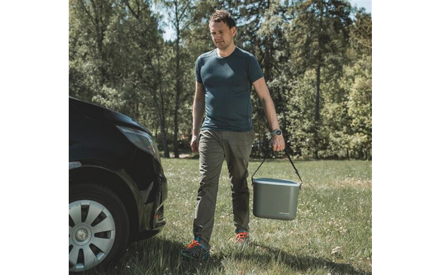 Homme marchant sur l'herbe près d'une voiture noire, portant une glacière gris-vert par la poignée ; arbres en arrière-plan