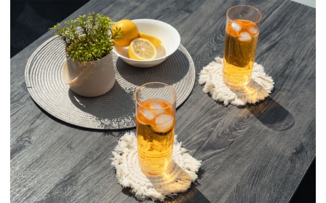 Zwei hohe Gläser mit orangefarbener Flüssigkeit, Eiswürfeln und Limettenscheiben auf hellen, gefransten Untersetzern auf einem dunklen Holztisch; daneben eine runde graue Tischmatte mit einer weißen Schale mit Zitronenscheiben und einer Topfpflanze