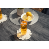 Glas mit bernsteinfarbener Flüssigkeit und Eiswürfeln auf weißem Makramee-Untersetzer auf dunklem Holztisch; weiße Schale mit ganzen und halbierten Zitronen und kleiner weißer Blumentopf im Hintergrund; links teilweise ein weiteres Glas sichtbar