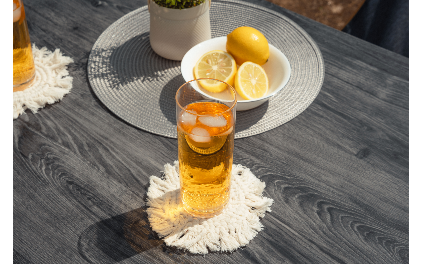 Glas mit bernsteinfarbener Flüssigkeit und Eiswürfeln auf weißem Makramee-Untersetzer auf dunklem Holztisch; weiße Schale mit ganzen und halbierten Zitronen und kleiner weißer Blumentopf im Hintergrund; links teilweise ein weiteres Glas sichtbar