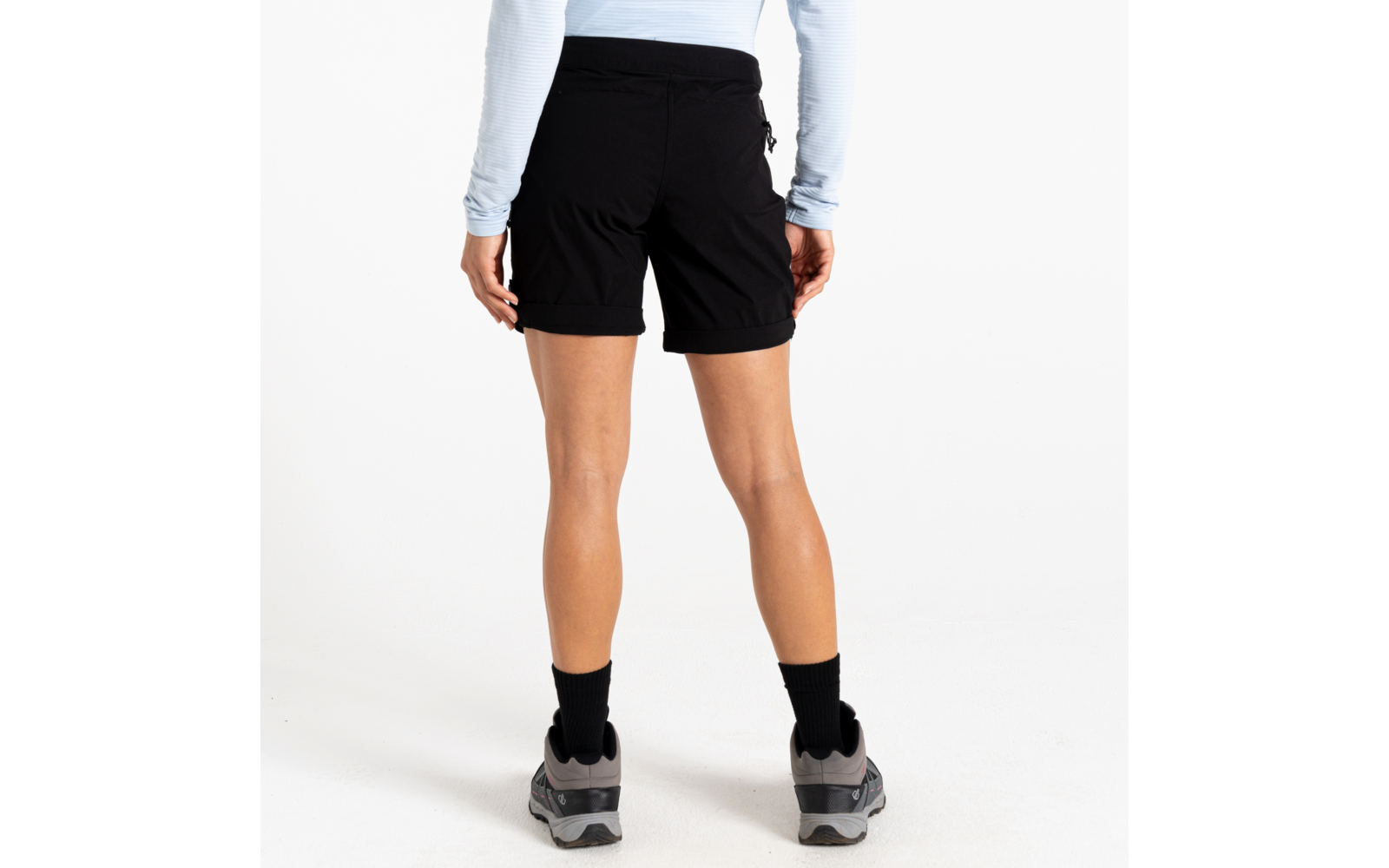 Achteraanzicht van een persoon vanaf de taille omlaag, zwarte shorts, licht langarmshirt, zwarte sokken en grijze sneakers op witte achtergrond