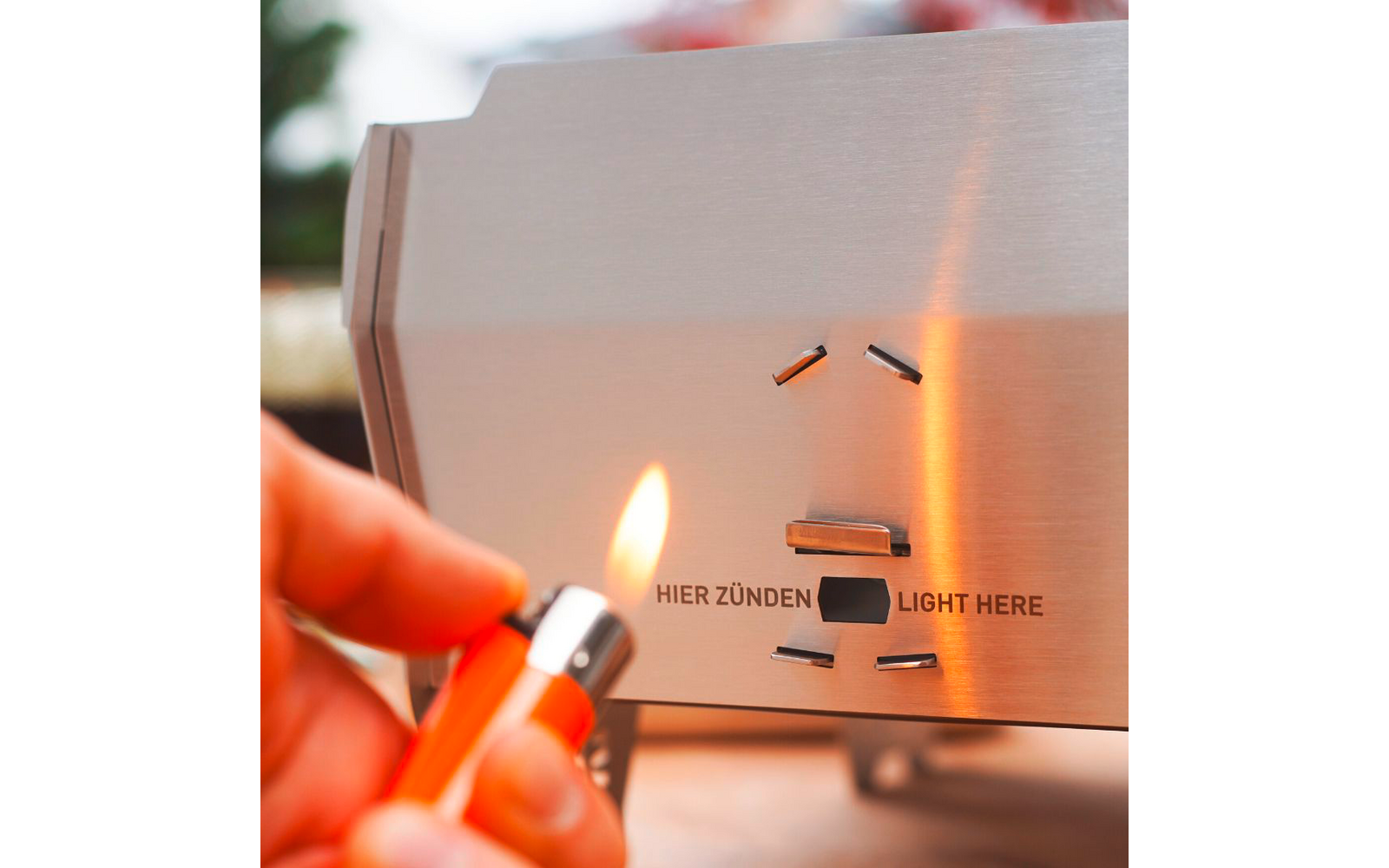 Edelstahlplatte mit der Aufschrift "HIER ZÜNDEN" und "LIGHT HERE", Hand hält orangenes Feuerzeug, kleine Flamme an einer Öffnung