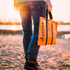 Untere Körperhälfte einer Person in Jeans und Sneakern am Sandstrand, in der Hand eine orangefarbene Tasche mit schwarzem Schriftzug 'SKOTTI'