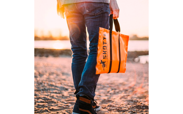 Untere Körperhälfte einer Person in Jeans und Sneakern am Sandstrand, in der Hand eine orangefarbene Tasche mit schwarzem Schriftzug 'SKOTTI'
