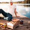 Mann mit weißer Mütze und T-Shirt sitzt auf einem Holzsteg am See; auf dem Steg ein kleiner Gaskocher mit Grill, eine Metallbox mit Speisen und ein Schneidebrett mit Kräutern