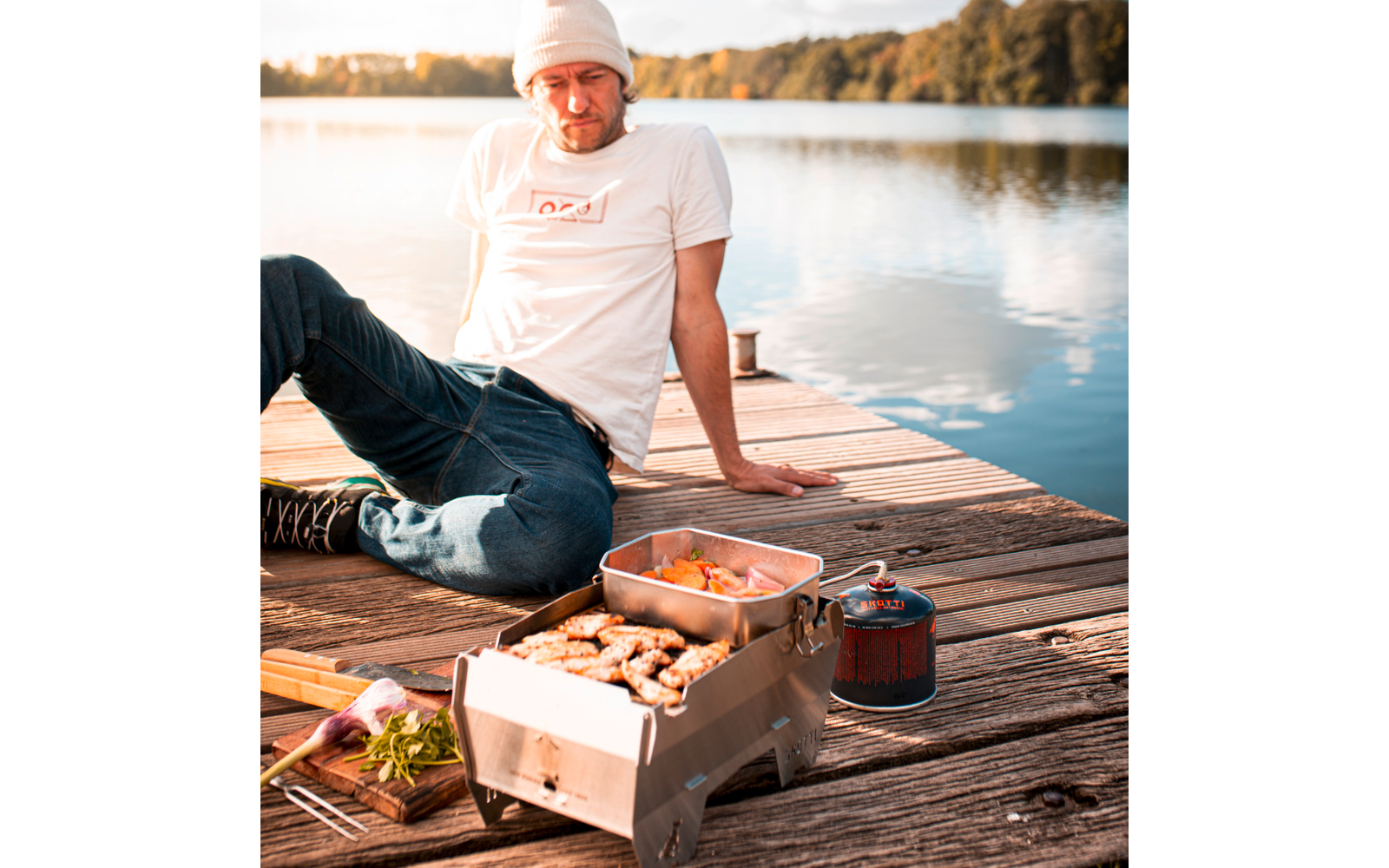 Mann mit weißer Mütze und T-Shirt sitzt auf einem Holzsteg am See; auf dem Steg ein kleiner Gaskocher mit Grill, eine Metallbox mit Speisen und ein Schneidebrett mit Kräutern