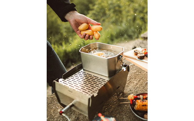 Hand hält mehrere kleine gelbe Kartoffeln über einen rechteckigen Metalltopf mit sichtbaren Wassertropfen auf einem Campingkocher; unscharfe Vegetation im Hintergrund und ein Teller mit gegrilltem Mais und Spießen am Rand