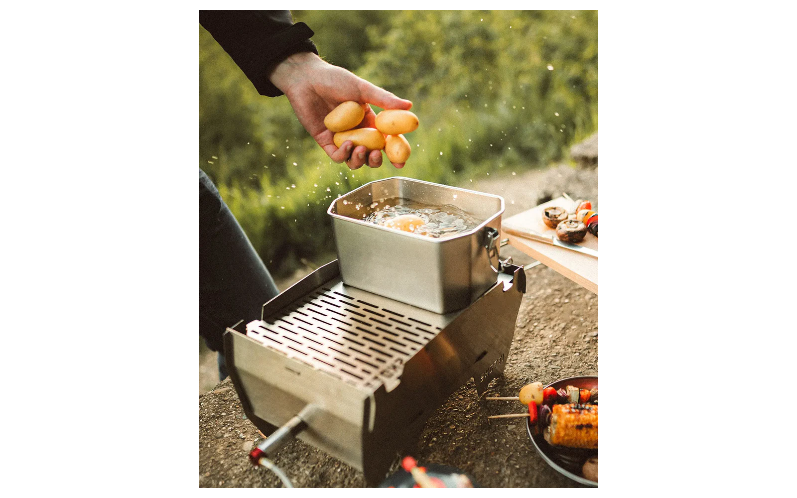 Hand hält mehrere kleine gelbe Kartoffeln über einen rechteckigen Metalltopf mit sichtbaren Wassertropfen auf einem Campingkocher; unscharfe Vegetation im Hintergrund und ein Teller mit gegrilltem Mais und Spießen am Rand