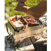 Tragbarer Metallgrill auf einem Holztisch mit Bratwürsten auf dem Rost, ein Metallpfännchen mit Spiegelei und Kirschtomaten, Hände über dem Grill