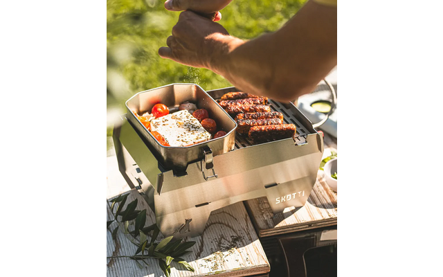 Tragbarer Metallgrill auf einem Holztisch mit Bratwürsten auf dem Rost, ein Metallpfännchen mit Spiegelei und Kirschtomaten, Hände über dem Grill