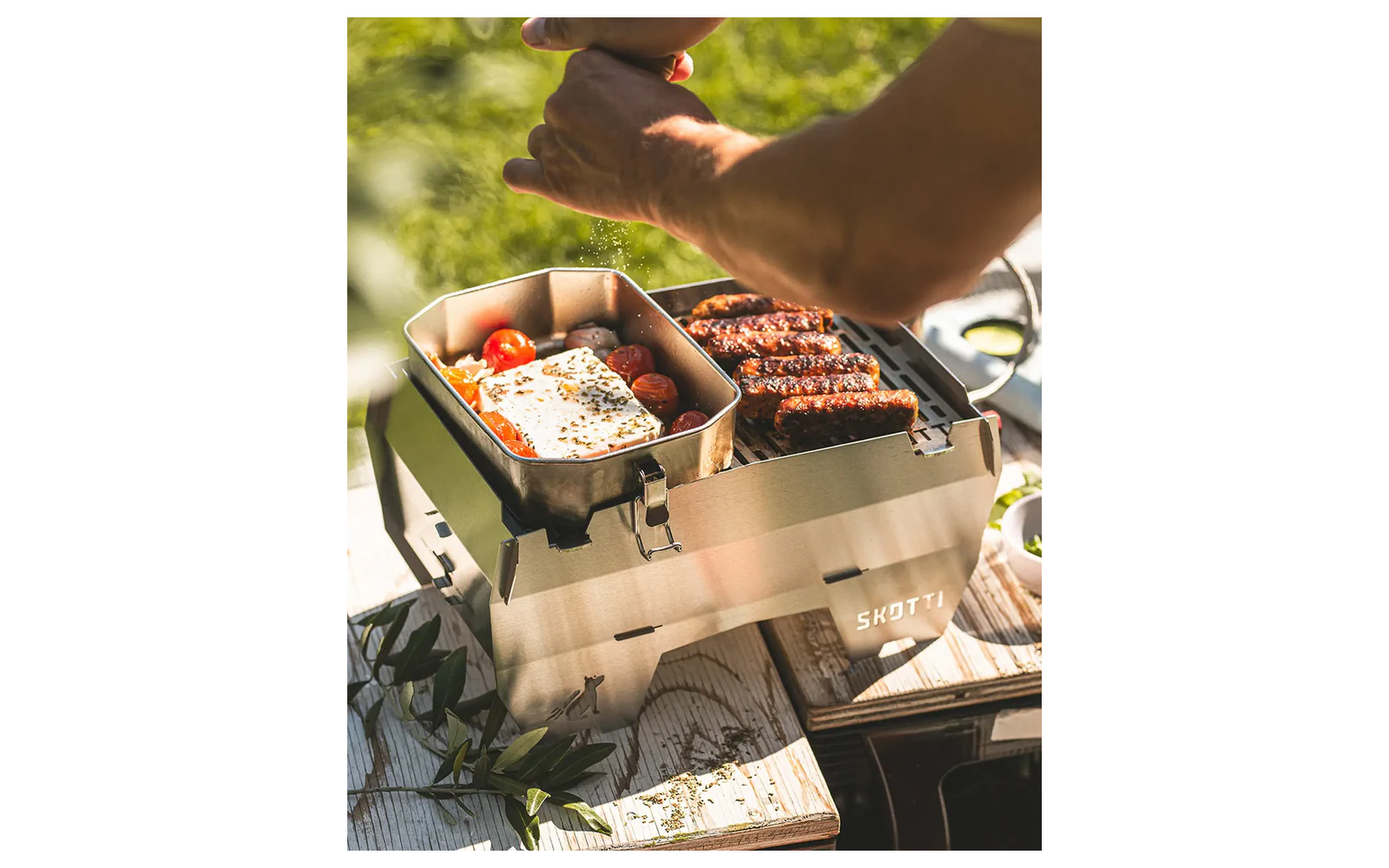 Tragbarer Metallgrill auf einem Holztisch mit Bratwürsten auf dem Rost, ein Metallpfännchen mit Spiegelei und Kirschtomaten, Hände über dem Grill