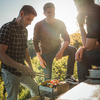 Drei Personen stehen um einen kleinen Grill im Freien, eine Person hält eine Grillzange, auf dem Grill liegen Spieße mit Gemüse und Fleisch, sonniger Hintergrund mit Bäumen