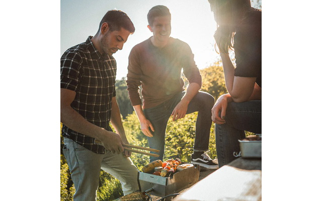 Drei Personen stehen um einen kleinen Grill im Freien, eine Person hält eine Grillzange, auf dem Grill liegen Spieße mit Gemüse und Fleisch, sonniger Hintergrund mit Bäumen