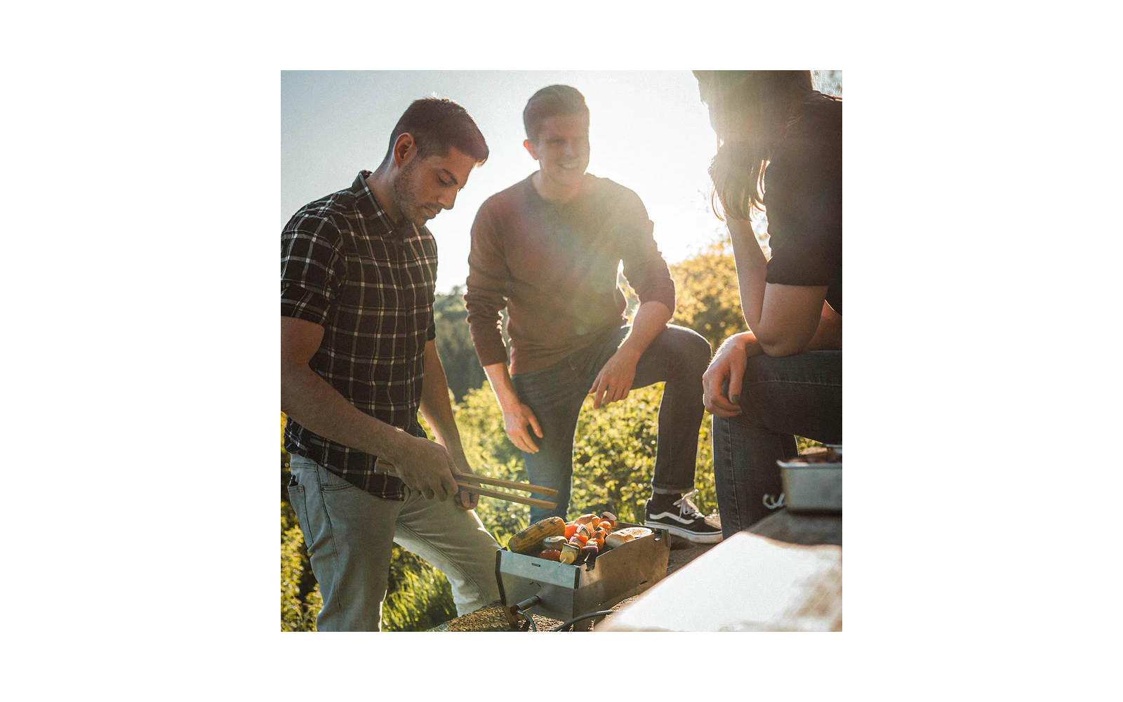 Drei Personen stehen um einen kleinen Grill im Freien, eine Person hält eine Grillzange, auf dem Grill liegen Spieße mit Gemüse und Fleisch, sonniger Hintergrund mit Bäumen