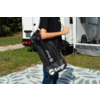 Torso de una persona con camiseta negra que lleva una bolsa negra con el logo 'Berger'; caravana, hierba y una alfombra estampada en el fondo