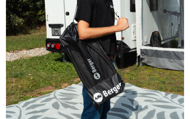 Torso de una persona con camiseta negra que lleva una bolsa negra con el logo 'Berger'; caravana, hierba y una alfombra estampada en el fondo